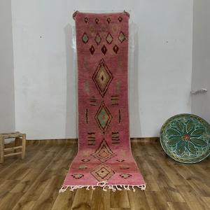 May include: A pink and red Moroccan rug with a diamond pattern. The rug is hanging on a white wall and has fringe on the bottom.