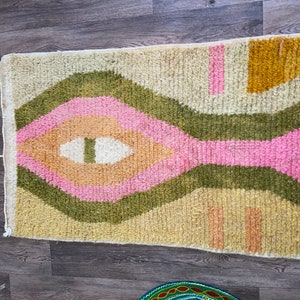 May include: A rectangular area rug featuring a high-pile texture with a cream-colored base. The design includes concentric diamond shapes in olive green, soft pink, and peach, accented by mustard yellow blocks and small pink and peach rectangular shapes.