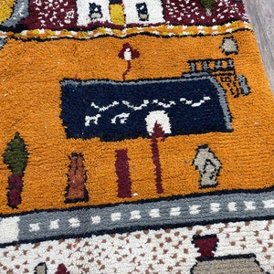 May include: A colourful rug with a geometric pattern and a depiction of a building with a blue roof. The rug has a variety of colours, including orange, red, blue, and green.