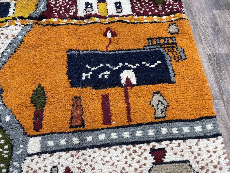 May include: A colourful rug with a geometric pattern and a depiction of a building with a blue roof. The rug has a variety of colours, including orange, red, blue, and green.