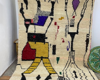 May include: Cream-colored, hand-woven rug with a geometric design. The rug features black, yellow, red, blue, and green shapes. The rug has a fringe along the bottom edge. The rug is approximately 6 feet by 4 feet.
