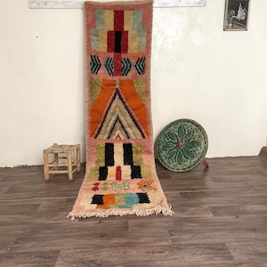 Op de afbeelding: Een lang, rechthoekig tapijt met een geometrisch patroon in tinten oranje, roze, groen, blauw en zwart. Het tapijt hangt aan een witte muur met een houten stok.