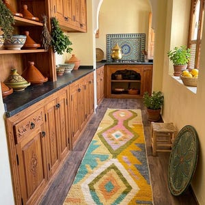 May include: A long rectangular area rug featuring a geometric pattern in yellow, pink, teal, and white with tasseled ends. The rug is placed on a dark wood floor in a kitchen with wooden cabinets, various ceramic tajines, and a tiled backsplash.