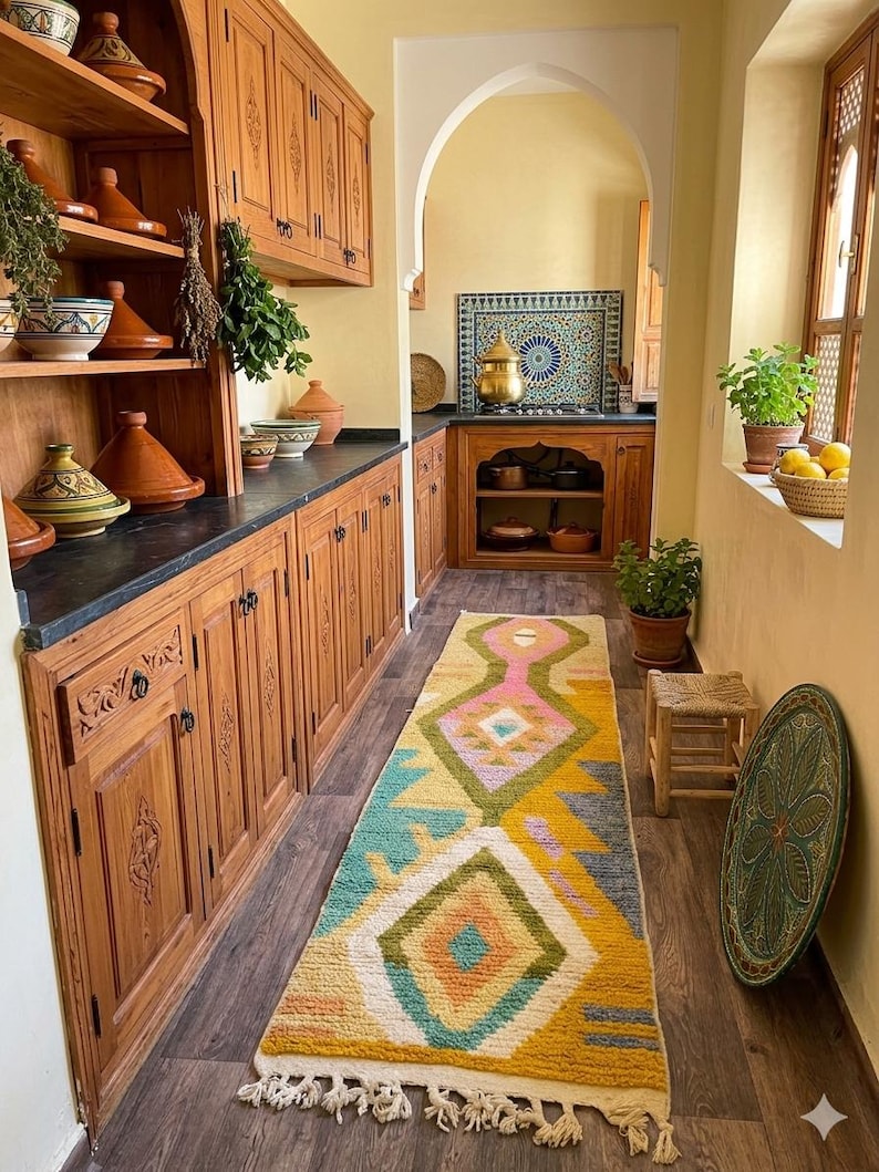 May include: A long rectangular area rug featuring a geometric pattern in yellow, pink, teal, and white with tasseled ends. The rug is placed on a dark wood floor in a kitchen with wooden cabinets, various ceramic tajines, and a tiled backsplash.