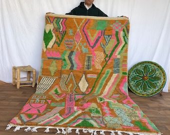 Puede incluir: Una alfombra marroquí grande y colorida con patrones geométricos en tonos de rosa, verde, marrón y blanco. La alfombra tiene un borde de flecos y está siendo sostenida por una persona.