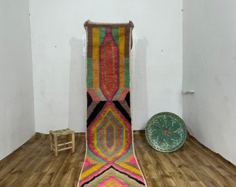 May include: A long, colourful rug with a geometric pattern in shades of pink, yellow, green, brown, and black. The rug has fringe on the bottom and is hanging on a white wall.
