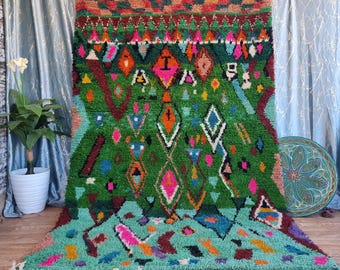 Puede incluir: Una alfombra marroquí colorida con un patrón geométrico en tonos de verde, rosa, azul y naranja. La alfombra tiene un borde de flecos y está colgada en una pared.