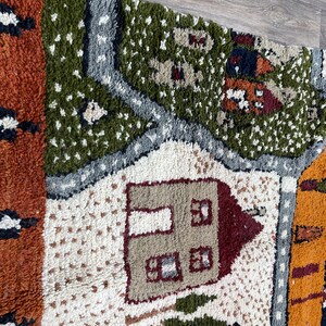 May include: A colourful, hand-woven rug with a whimsical design featuring houses, trees, and other details. The rug is made of wool and has a textured surface.