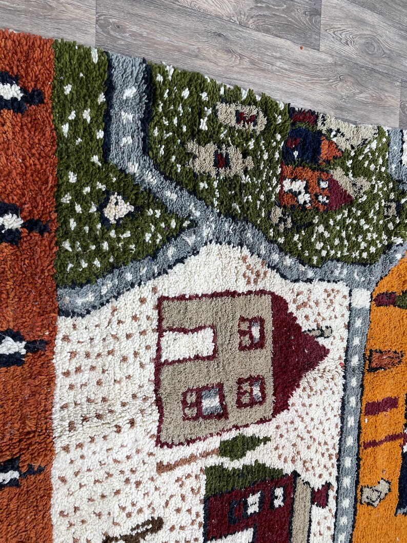 May include: A colourful, hand-woven rug with a whimsical design featuring houses, trees, and other details. The rug is made of wool and has a textured surface.