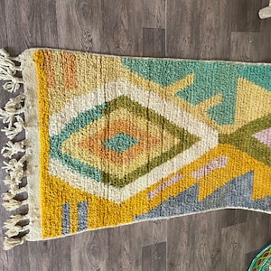 May include: A rectangular area rug featuring a high-pile texture with a geometric pattern of diamonds and lines. The color palette includes mustard yellow, sage green, cream, soft orange, and muted blue, finished with cream-colored fringe tassels.