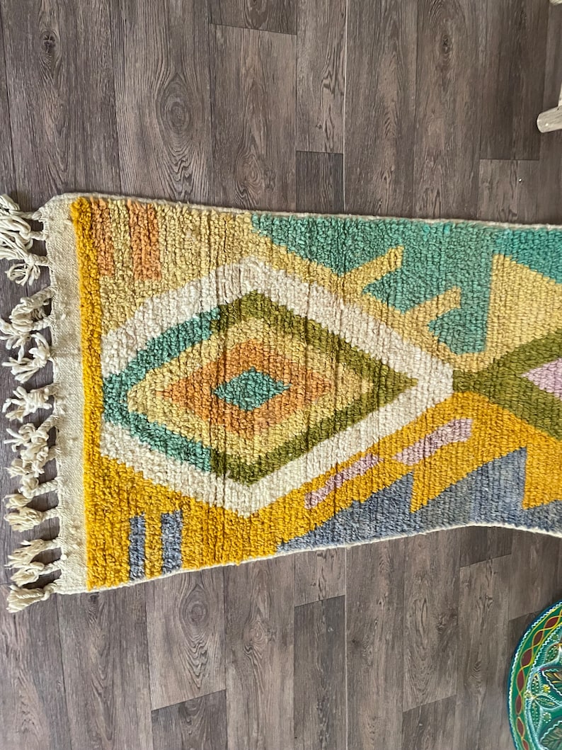 May include: A rectangular area rug featuring a high-pile texture with a geometric pattern of diamonds and lines. The color palette includes mustard yellow, sage green, cream, soft orange, and muted blue, finished with cream-colored fringe tassels.