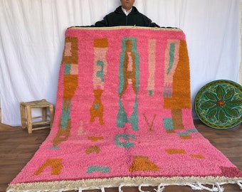 May include: A pink Moroccan rug with abstract geometric patterns in shades of orange, teal, and brown. The rug has a fringe border and is being held up by a person.