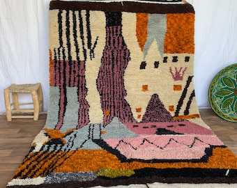 May include: A large, rectangular, hand-woven rug with a colorful, abstract design. The rug features a cream-colored background with geometric shapes in orange, pink, black, and gray. The rug has a fringe along the bottom edge.