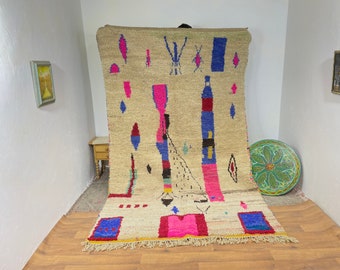 May include: A large, hand-woven rug with a beige background and colorful geometric patterns in shades of pink, blue, and red. The rug has a fringe border and is displayed on a wooden floor.