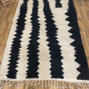 May include: Black and white striped rug with a geometric pattern. The rug has a fringe border and is made of a soft, fluffy material.