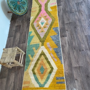 May include: A long, rectangular runner rug featuring a cream-colored base with a geometric pattern in shades of mustard yellow, olive green, dusty pink, teal, and slate blue. The design includes large diamond shapes and fringe at the ends.