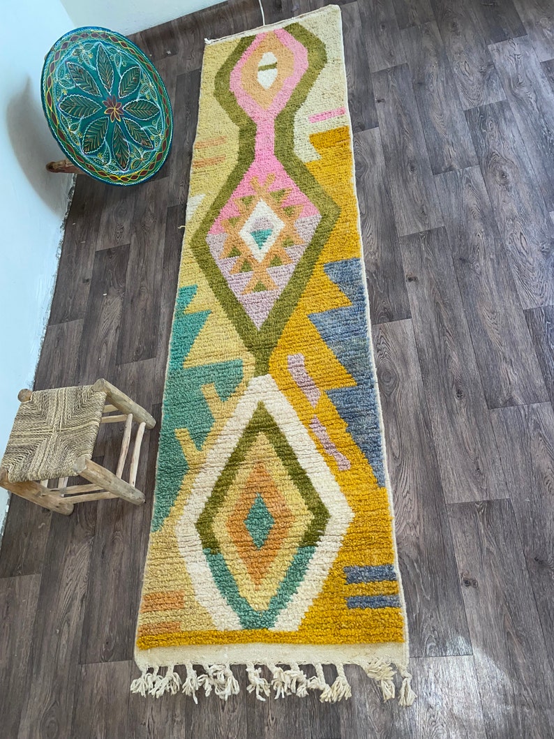May include: A long, rectangular runner rug featuring a cream-colored base with a geometric pattern in shades of mustard yellow, olive green, dusty pink, teal, and slate blue. The design includes large diamond shapes and fringe at the ends.