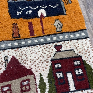 May include: A colourful rug with a whimsical design featuring houses, trees, and a black and white sign. The rug is made of a soft, textured material and has a variety of colours, including red, orange, yellow, green, and blue.