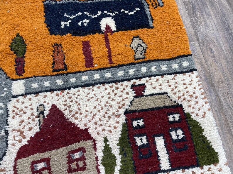 May include: A colourful rug with a whimsical design featuring houses, trees, and a black and white sign. The rug is made of a soft, textured material and has a variety of colours, including red, orange, yellow, green, and blue.