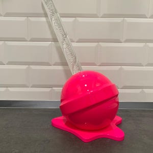 May include: A bright pink lollipop sculpture with a clear acrylic stick. The lollipop is melting and dripping onto a dark surface.
