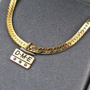 Puede incluir: Un collar de cadena de oro con un colgante que dice "D-M-E" en letras negras y tiene tres pequeñas piedras preciosas rojas.