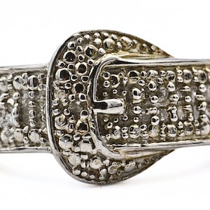 Könnte beinhalten: Ein silberfarbener Ring in Form einer Gürtelschnalle. Der Ring hat eine strukturierte Oberfläche mit kleinen, erhabenen Details. Das Schnallendesign umfasst einen Mittelsteg und einen Rahmen, alles in glänzendem Silber.