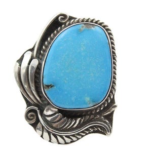 Op de afbeelding: Een sierlijke zilveren ring met een opvallende, ovale turkoois edelsteen. Het ontwerp van de ring omvat ingewikkelde blad- en wervelpatronen, waardoor een gedurfd en decoratief sieraad ontstaat. De turkoois steen is levendig blauw.