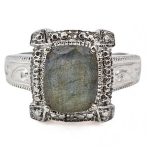 May include: A silver ring featuring a large, rectangular, gray-green gemstone. The ring has a detailed, textured setting with small embellishments and decorative patterns on the band. The gemstone is the focal point, showcasing a subtle play of colors.