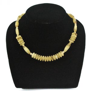 Könnte beinhalten: Eine Halskette aus hellbraunen ovalen und runden Perlen. Die Halskette befindet sich auf einem schwarzen Samt-Präsentationsständer.