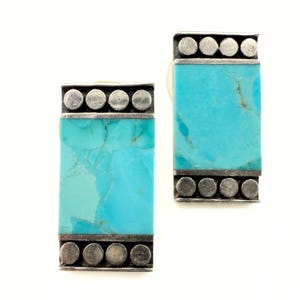Può includere: Un paio di orecchini rettangolari in turchese con una montatura in metallo color argento. Le pietre turchesi presentano venature naturali. La parte superiore e inferiore di ogni orecchino presenta una fila di piccoli accenti metallici rotondi.