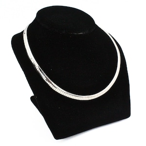 May include: A silver-colored, flat snake chain necklace displayed on a black velvet jewelry bust. The necklace has a smooth, reflective surface and a flexible design. The jewelry bust is set against a white background.