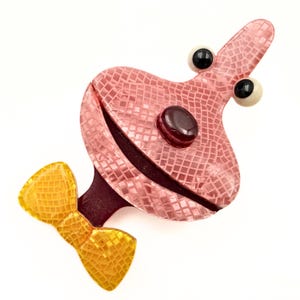 May include: A pink brooch shaped like a stylized character with a textured grid pattern, featuring two protruding eyes with black pupils, a dark red oval nose, and a wide mouth. A bright yellow bow tie with a grid texture is attached at the base.