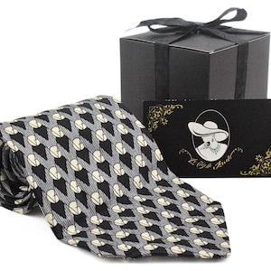 May include: A rolled black, gray, and cream-colored tie with a repeating geometric pattern. A black gift box with a bow and a business card with a woman's silhouette are behind the tie. The business card has the text "Le Belle Aurel".