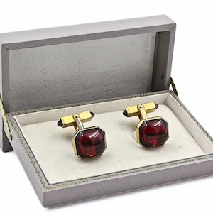 May include: A pair of gold-toned cufflinks featuring faceted, deep red, octagonal-shaped stones. The cufflinks are presented inside a rectangular, grey-colored storage box with a white interior lining and a hinged lid that is open.
