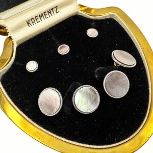 Può includere: Un set vintage di gemelli e bottoni da camicia KREMENTZ in madreperla in una scatola di presentazione con bordi dorati. Il set include pezzi rotondi e ovali con superfici iridescenti, su un interno in velluto nero.