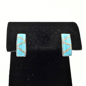 May include: A pair of rectangular turquoise earrings with silver-colored borders. The earrings feature an inlaid design of diagonal lines in a lighter color, possibly mother-of-pearl or a similar iridescent material. The earrings are displayed on a black surface.