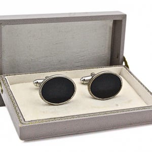 May include: A pair of oval-shaped cufflinks with a smooth black center and a silver-toned metal border, presented inside a rectangular gray hinged storage box with a cream-colored interior lining and a decorative stitched edge detail.