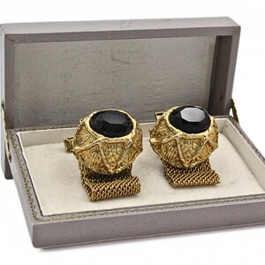 May include: A pair of gold-toned cufflinks with large, black, oval-shaped stones, presented in a gray box with a cream-colored interior. The cufflinks have a textured design and are connected by a gold chain.