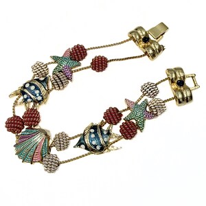 Könnte beinhalten: Ein goldfarbenes Charm-Armband mit zwei Kettensträngen und emaillierten Meeresmotiven wie Fischen, einer Muschel und einem Seestern. Das Armband ist mit kleinen, texturierten roten und weißen kugelförmigen Perlen verziert und hat einen Metallverschluss.