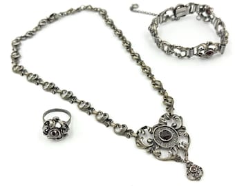 May include: A silver-coloured jewellery set comprising a necklace, bracelet, and ring. The necklace features a central pendant with a dark red stone. The bracelet and ring also have dark red stones. The set has an ornate, floral design.