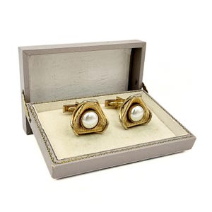 May include: A pair of gold-toned cufflinks featuring a triangular shape with a textured, ribbed surface and a central white pearl-like cabochon. The cufflinks are presented inside a rectangular, light gray hinged box with a cream-colored interior.