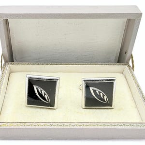 Puede incluir: Un par de gemelos cuadrados con un centro negro y un diseño de hoja plateada, presentados en una caja gris claro con un interior color crema. Los gemelos se muestran abiertos, mostrando el diseño.