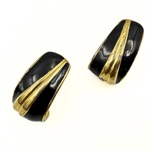 May include: A pair of vintage clip-on earrings. Each earring features a black enamel finish with a gold-tone metal accent. The earrings have a curved, rectangular shape with a diagonal stripe design.