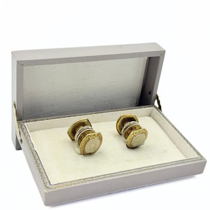 May include: A pair of gold and silver cufflinks with a geometric design, presented in a gray box with a cream-colored interior. The cufflinks have a textured, layered appearance, and the box is open.