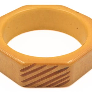 May include: A vintage, octagonal bangle bracelet in a warm, amber hue. The bracelet features a series of diagonal, brown stripes on one of its facets. The design is simple and geometric, suitable for everyday wear. This piece is a classic jewelry item.