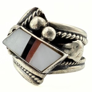 May include: A silver ring with a geometric design. The ring features a central diamond-shaped inlay with white, black, and reddish-brown sections. The band has a textured, rope-like design and small silver beads. The ring has a vintage or Native American aesthetic.