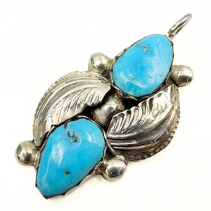 May include: A silver pendant featuring two teardrop-shaped turquoise stones set in polished metal. The design includes embossed leaf motifs on the sides, small spherical silver accents at the corners, and a metal bail at the top for hanging.