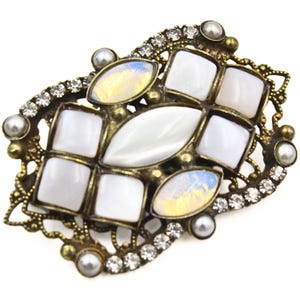 May include: An antique-style brooch with a bronze-toned, filigree frame. It features white, square, and oval-shaped stones, iridescent highlights, faux pearls, and clear rhinestones. A classic accessory for any outfit.