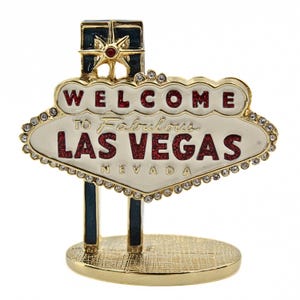 May include: Gold-colored metal replica of the iconic "Welcome to Fabulous Las Vegas" sign with red glitter lettering and a white background. The sign is decorated with clear rhinestones and sits on a gold-colored base.
