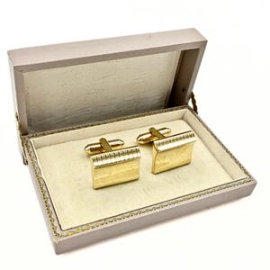 May include: A pair of gold-toned cufflinks with a rectangular design, presented in a light gray box with a cream-colored interior. The cufflinks have a textured, ridged detail on the front.